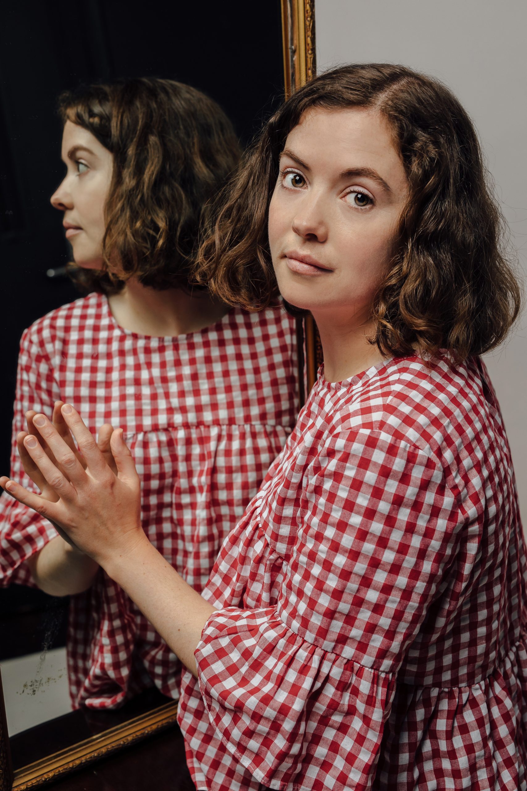 Clare as an adult reflected in a mirror wearing a red and white gingham top