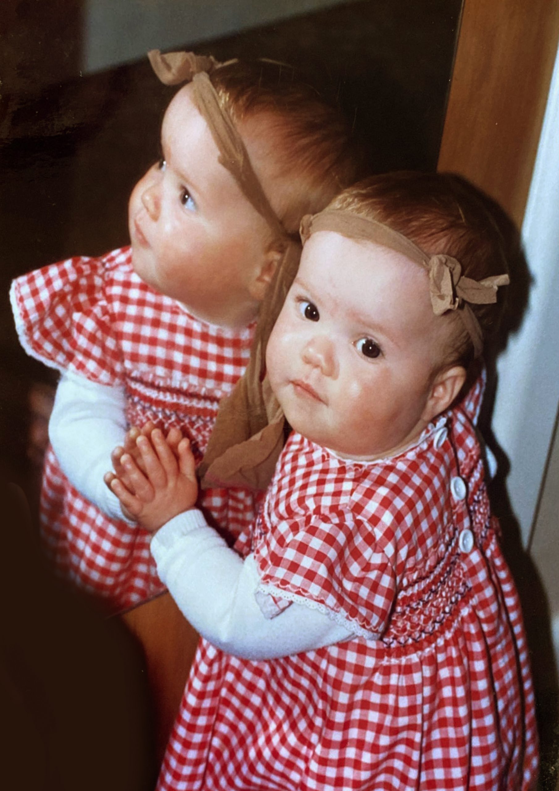 Clare as a baby reflected in a mirror wearing a red and white gingham top