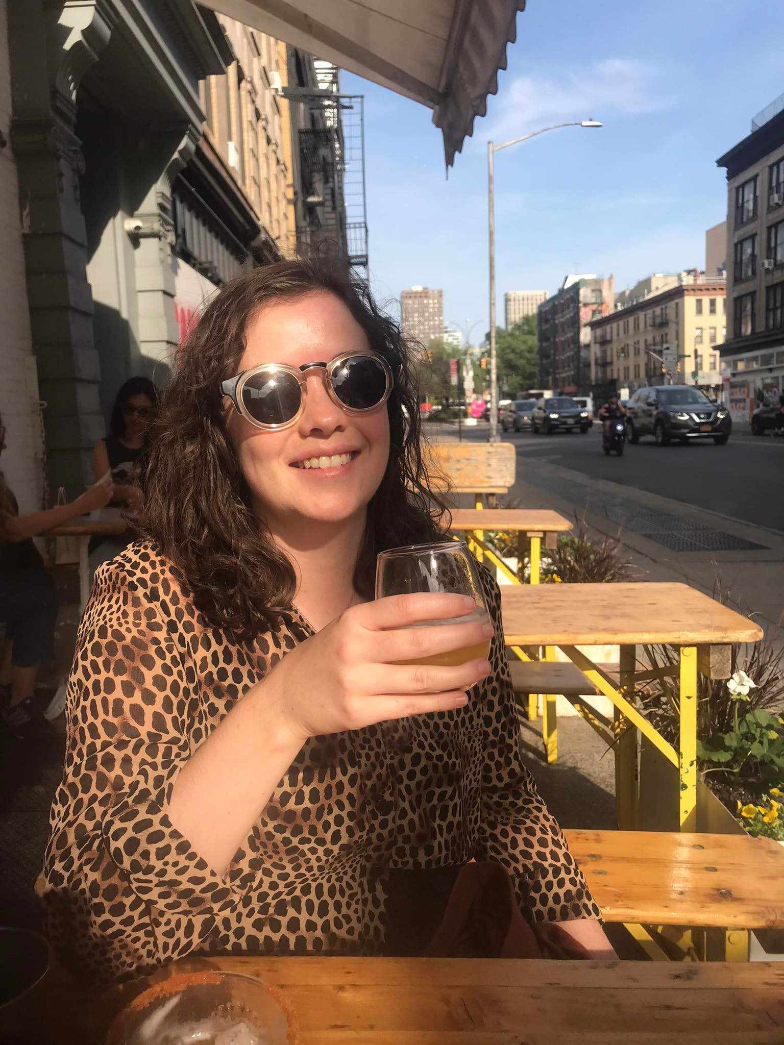 Clare wears dark round sunglasses and aleaopard print shirts and sits smiling in the sunshine, a drink in her hand