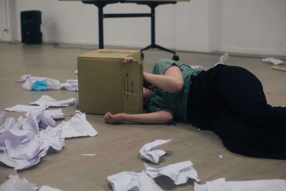 Clare lying down with her head in a cardboard box, papers strewn on the ground